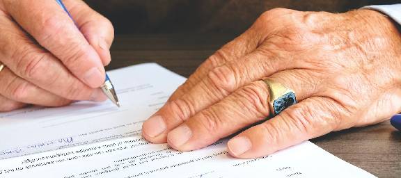 Older caucasian female hands signing a document with a blue ballpoint pen, hands adorned with a gold ring with opal inset on the left-hand ring-finger and a white metal loop on the right-hand ring-finger.
